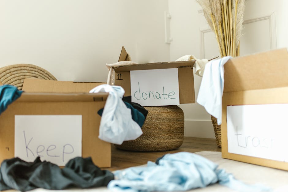 A room with three large cardboard boxes labeled 'Keep', 'Donate', and 'Trash', placed on a wooden floor near a woven basket and a window with floral curtains. The boxes contain various clothing items, some draped over the edges and partially visible. The scene is well-lit with natural daylight, emphasizing the cleanliness and organization within the space. This setup reflects a decluttering process with an emphasis on maintaining hygiene and order, consistent with the eco-friendly cleaning services provided by Hounslow Cleaners for domestic cleaning and surface sanitisation in residential settings.