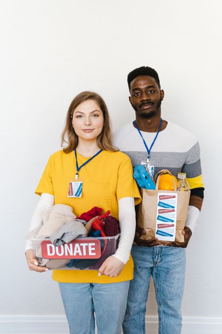A young woman and man standing side by side against a plain white wall, both wearing yellow and grey shirts with blue lanyards and badges. They are holding donation boxes; the woman's box is labeled 'DONATE' and contains clothes, while the man's box contains toiletries and household items. The setting suggests a community or charity event related to local support and donations, and the image conveys a positive, cooperative atmosphere associated with community engagement.
