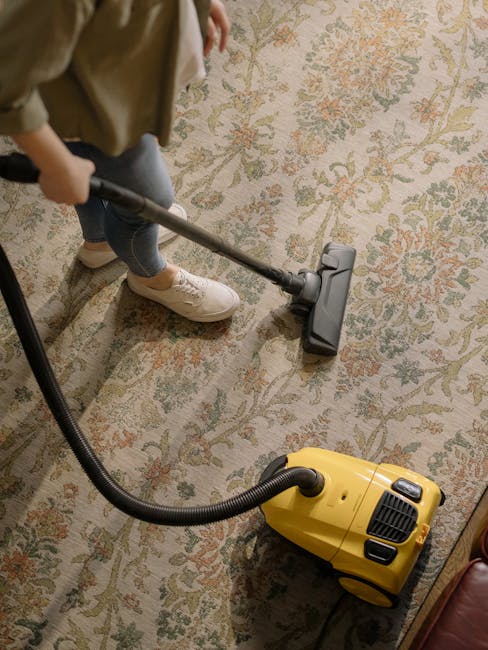 A child dressed in casual clothing and white sneakers is vacuuming an oriental-style area rug with a patterned floral design in a room with soft lighting. The vacuum cleaner, which appears to be a canister model with a yellow body and black hose, is moving across the plush, textured surface. The room features a wooden floor partially visible beneath the rug, and the surrounding furniture includes a glimpse of a leather armchair or couch in the corner. The scene demonstrates surface cleaning as part of a domestic cleaning process, with Hounslow Cleaners' professional services focusing on maintaining hygiene and deep cleaning. The image emphasizes thorough vacuuming to remove dust and improve cleanliness on woven, fibrous materials.
