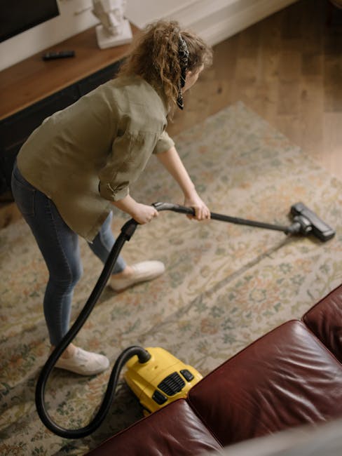 A woman with curly hair, wearing a beige jacket, dark jeans, and white shoes, is using a yellow vacuum cleaner to deep clean a patterned area rug in a living room. She is holding the vacuum's hose nozzle and working on the surface of the floral-patterned carpet, which appears clean and well-maintained. The room features wooden flooring adjacent to the rug, with a dark brown leather sofa visible in the foreground, and a wooden sideboard along the wall in the background, set under neutral-colored walls. Natural light illuminates the space, highlighting the polished surfaces and tidy appearance. Hounslow Cleaners provides professional domestic cleaning services that include surface cleaning and sanitisation to maintain hygiene and cleanliness in residential settings, as reflected in the focus on vacuuming and deep cleaning processes shown here.