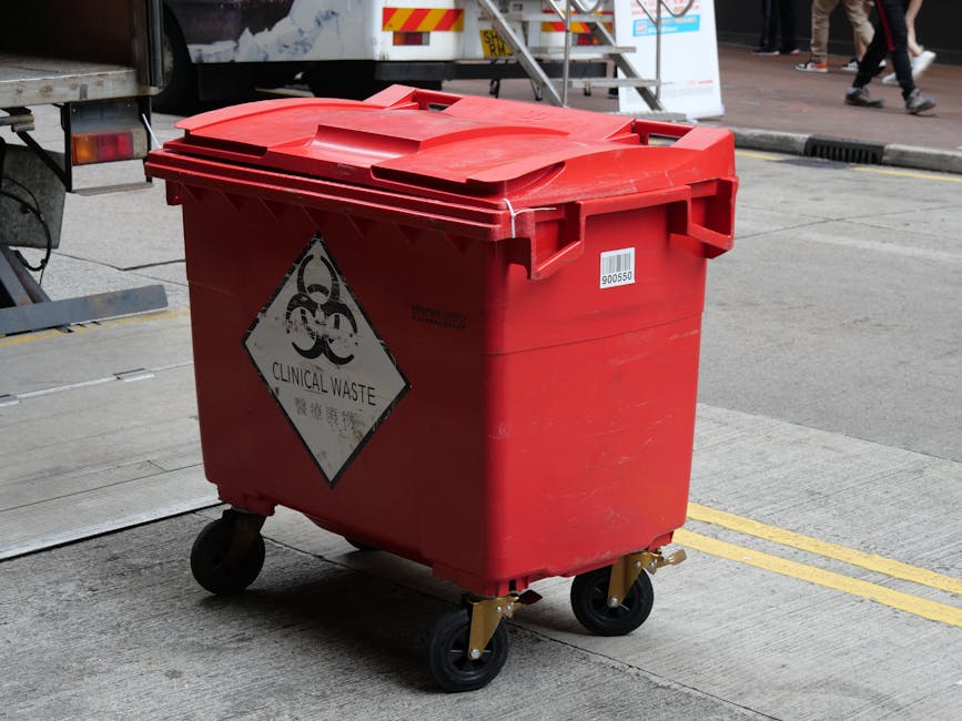 A red clinical waste bin with a black biohazard and 'Clinical Waste' label, situated on a concrete sidewalk in an urban area. The bin has a closed lid and four black wheels, with some visible dirt and smudges on its surface. In the background, there is part of a truck and pedestrians walking on the street. The scene is well-lit with natural daylight, emphasizing the clean condition of the surroundings. This image is associated with Hounslow Cleaners' service page on hazardous waste disposal and cleaning, demonstrating proper handling of clinical waste containers as per Hounslow Council regulations.
