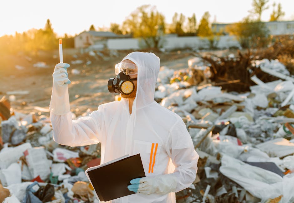 A person dressed in full protective gear, including a white disposable suit, gloves, goggles, and a respirator mask, standing amidst a large outdoor dump site filled with scattered waste, trash bags, and debris. The individual is holding a clipboard in one hand and a small test tube or sample container in the other, inspecting the area under natural sunlight. The surrounding environment is rugged, with mixed materials like plastic, paper, and discarded objects, indicating a hazardous waste site requiring careful assessment and management. This scene emphasizes safety protocols and the importance of thorough site inspection for hazardous waste disposal, aligning with the guidelines discussed on Hounslow Council rules for hazardous waste disposal and cleaning. Hounslow Cleaners advocates for professional cleaning and sanitisation practices, especially in environments with potential health risks.
