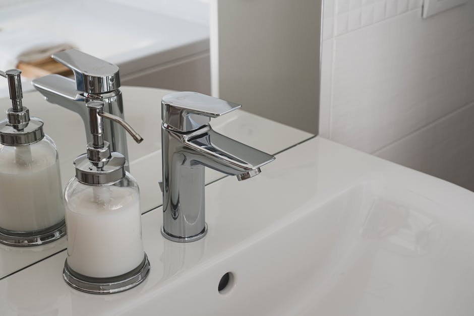 Close-up view of a white bathroom sink with a modern chrome faucet, accompanied by two clear glass soap dispensers filled with white liquid soap and lotion, positioned on the sink's surface. The surface appears clean and shiny, reflecting light, with a clean mirror behind the sink and tiled wall in the background, indicating a well-maintained domestic space. The arrangement exemplifies surface cleaning and hygiene practices, consistent with professional domestic cleaning services by Hounslow Cleaners for end of tenancy and general bathroom upkeep.