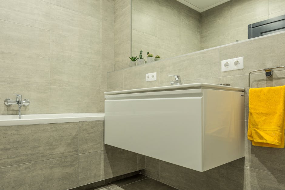 The image depicts a modern bathroom with beige ceramic wall and floor tiles, showcasing a clean and well-maintained environment. On the left, there is a white bathtub with a chrome faucet and fixtures. To the right, a white wall-mounted vanity unit with a smooth, glossy surface and integrated sink is visible, topped by a chrome single-handle faucet. Above the vanity, there are two electrical outlets and a large mirror reflecting light, enhancing the bright ambiance. A small ledge behind the sink holds decorative potted plants, adding a touch of greenery. Adjacent to the vanity, a chrome towel rail holds a neatly folded yellow towel. The overall scene emphasizes surface cleaning, hygiene, and deep cleaning processes, consistent with domestic cleaning services provided by Hounslow Cleaners as highlighted on the Hounslow end of tenancy cleaning Bath Road specialists page.