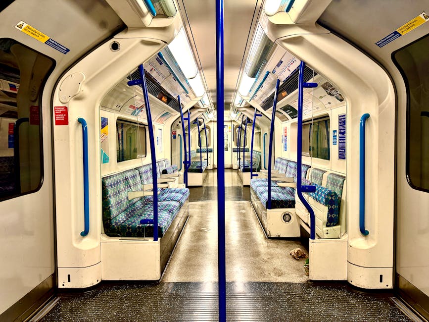 Interior of an empty train carriage featuring clean, polished surfaces with a predominantly white and blue color scheme. The floor appears well-maintained and free of dirt, with textured black flooring near the entrance. The seating consists of upholstered benches with blue and green patterned fabric, positioned along the sides of the carriage. Blue handrails run vertically and horizontally for passenger stability. The windows are clear, allowing ambient light into the space, and the ceiling has integrated lighting fixtures. Visible signs with safety instructions are affixed to the walls, contributing to the tidy and hygienic environment. This image exemplifies professional surface cleaning and maintenance suitable for train carriage hygiene standards, as provided by Hounslow Cleaners.