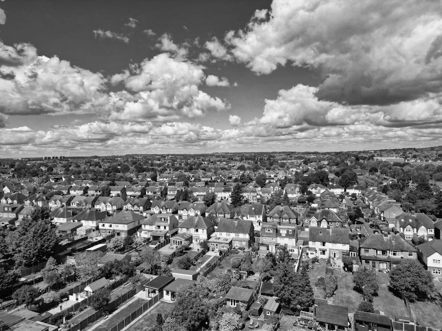 A black-and-white aerial photograph of a suburban residential area showing rows of detached and semi-detached houses with individual gardens, driveways, and rooftops made of various materials such as tiles and shingles. The streets are lined with trees and greenery, and the overall scene is illuminated by natural daylight under a sky filled with large, fluffy clouds. The image conveys a neat and orderly neighborhood landscape, with well-maintained properties typical of suburban housing. This visual may relate to domestic or community cleaning services provided by Hounslow Cleaners, highlighting the importance of surface cleaning and maintenance within residential environments.