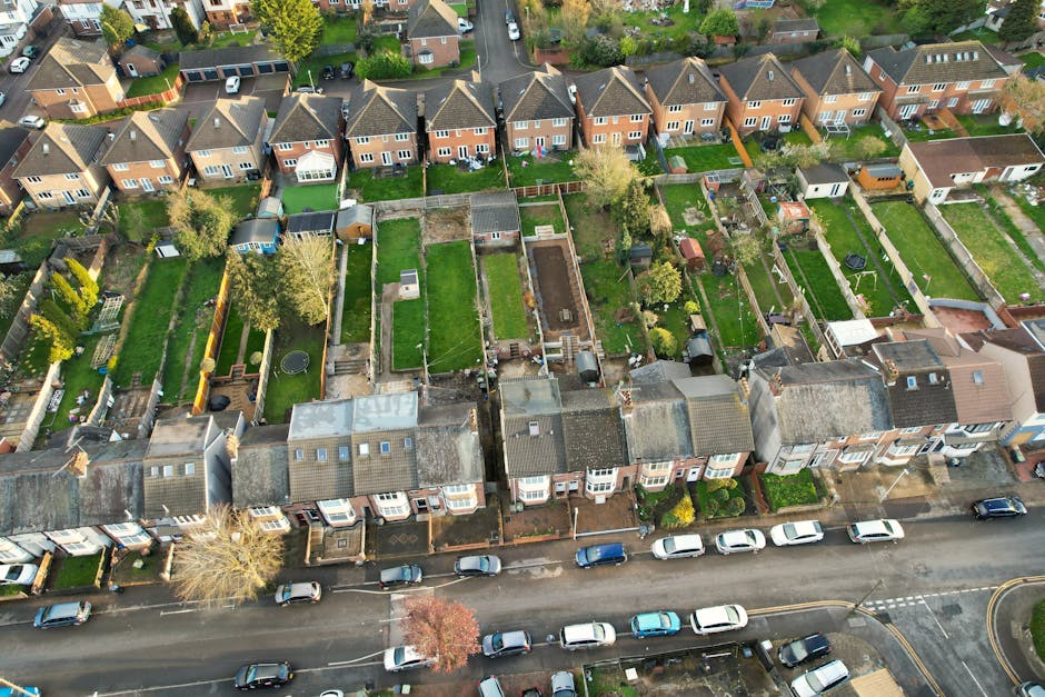 An aerial view of a residential street in Hounslow showcasing a row of terraced houses with various backyard sizes, many enclosed by wooden or brick fences. Some gardens feature well-maintained grass lawns, paved pathways, and small trees or shrubs. Several backyards include garden sheds, patios, or decking areas, with one yard having a trampoline. The street below is lined with parked cars along the curb, and surrounding greenery is visible in the distance. The lighting indicates a clear day with natural sunlight illuminating the scene. This detailed view highlights the typical suburban housing layout and outdoor spaces, exemplifying properties that could benefit from professional domestic cleaning services like those offered by Hounslow Cleaners.