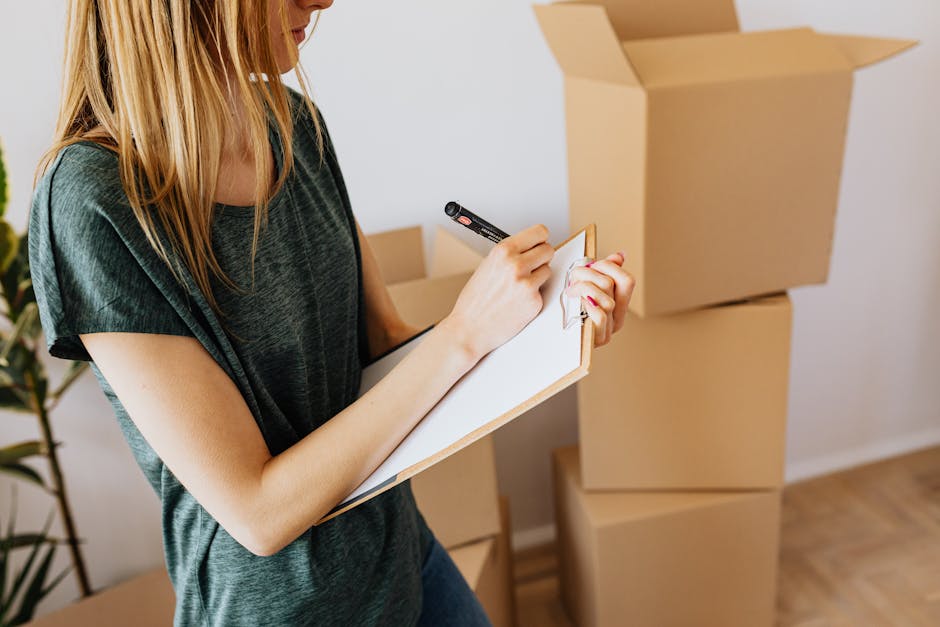 A woman with long blonde hair, wearing a dark green T-shirt, is writing on a white notepad with a black marker. She is standing in a room with several light brown cardboard boxes, some stacked on each other, indicating a moving or cleaning process. The room has a wooden floor and a white wall in the background. A potted plant with green leaves is partially visible on the left side of the image. The scene reflects an organized approach to end-of-tenancy cleaning, with the woman possibly creating a checklist for surface cleaning or sanitisation, which aligns with the landlord checklist Hounslow Council end of tenancy cleaning rules, HOUNSLOW. The overall setting emphasizes cleanliness and preparation for property inspection, consistent with services provided by Hounslow Cleaners in the cleaning category.
