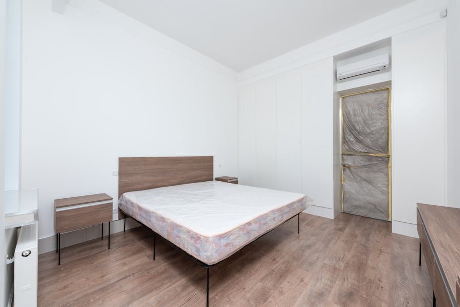 A minimalist bedroom featuring a double bed with a wooden headboard and a plain white mattress. There are two matching wooden nightstands, one on each side of the bed. The floor is covered with light-colored wood laminate, contributing to a clean and modern aesthetic. Against the white walls, there is a built-in wardrobe with sleek, handleless doors. An air conditioning unit is mounted high on the wall near an unadorned door covered with protective plastic and gold tape, indicating recent or ongoing renovation. The room is well-lit with natural light, highlighting the tidy, dust-free surfaces, and creating a fresh, hygienic environment suitable for domestic cleaning or surface sanitisation by Hounslow Cleaners.