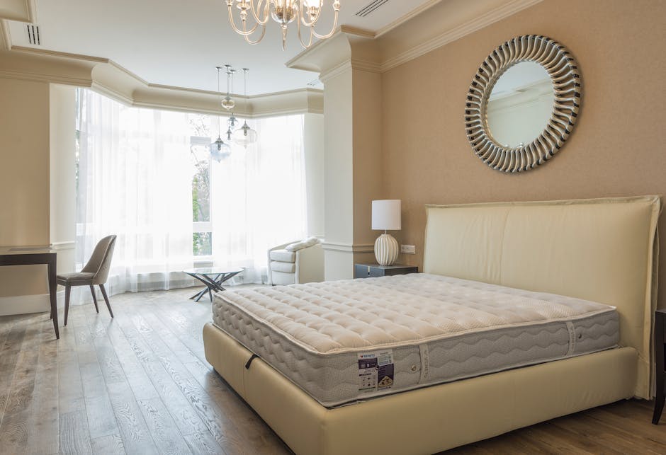 A bright, modern bedroom featuring a large bed with a white upholstered headboard and a clean, beige mattress. The room has a light-colored wooden floor and is illuminated by natural light coming through large, sheer white curtains covering a bay window. To the right of the bed, there is a dark grey bedside table with a white ceramic lamp. On the left side, a small black desk with a beige upholstered chair is positioned against the wall, with a framed mirror hanging above it. A white armchair is placed near the window, and a round glass coffee table with a metal base sits in front of the window. The room's décor includes a round, decorative mirror on the wall, and the space appears spotless and well-maintained, reflecting a professional domestic cleaning environment provided by Hounslow Cleaners.