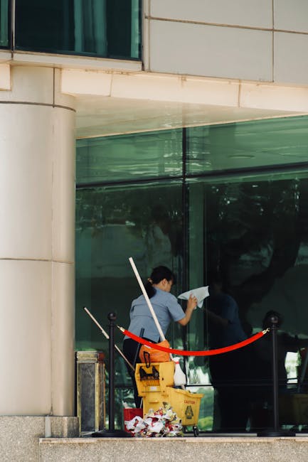 A professional cleaner from Hounslow Cleaners is seen outside a modern commercial building with large glass windows. The cleaner, dressed in casual work attire, is using a cloth to wipe the glass surf