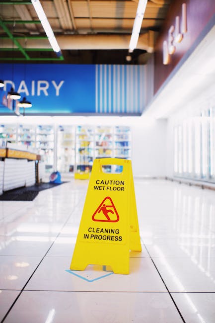 Inside a supermarket aisle, a bright yellow 'Caution Wet Floor' sign with black and red text and symbols is prominently displayed on the glossy white ceramic tiled floor, indicating ongoing cleaning and a potential slip hazard. The aisle features natural light streaming in from large windows on the right, illuminating the spacious area. To the left, a shelving unit stocked with products is visible, with some items arranged neatly. Overhead, modern lighting fixtures hang from the ceiling, which has a wooden finish. The clean, reflective surface of the tiles and the organized environment reflect professional cleaning standards. The signage and overall tidiness suggest that Hounslow Cleaners specializes in thorough surface cleaning and sanitisation, maintaining hygiene and safety in commercial premises such as this supermarket, aligning with their service of deep cleaning and surface sanitation for business premises in Hounslow.