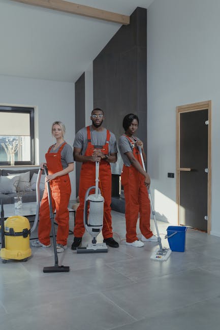 Three professional cleaning specialists from Hounslow Cleaners in orange overalls stand inside a modern living room, equipped with vacuum cleaners, mops, and cleaning buckets, ready to perform surface cleaning and deep cleaning tasks. The room features large windows allowing natural light, a sleek tiled floor, a grey accent wall, and contemporary furniture, including a sofa and coffee table. The team appears prepared to provide comprehensive sanitisation and maintenance for commercial or domestic premises, demonstrating attention to cleanliness and hygiene standards. The image emphasizes the professional approach of Hounslow Cleaners in maintaining spotless and well-maintained indoor environments.